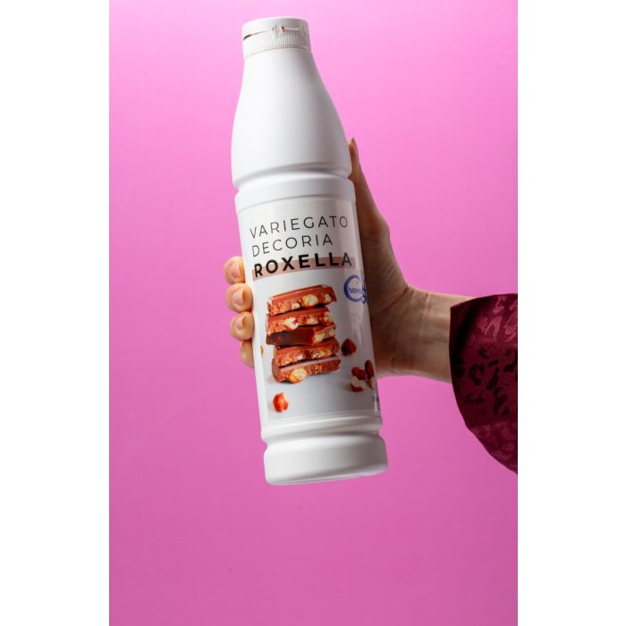 Hand holding a white bottle labeled "Variegato Decoria Roxella" with an image of chocolate and hazelnuts, against a pink background.