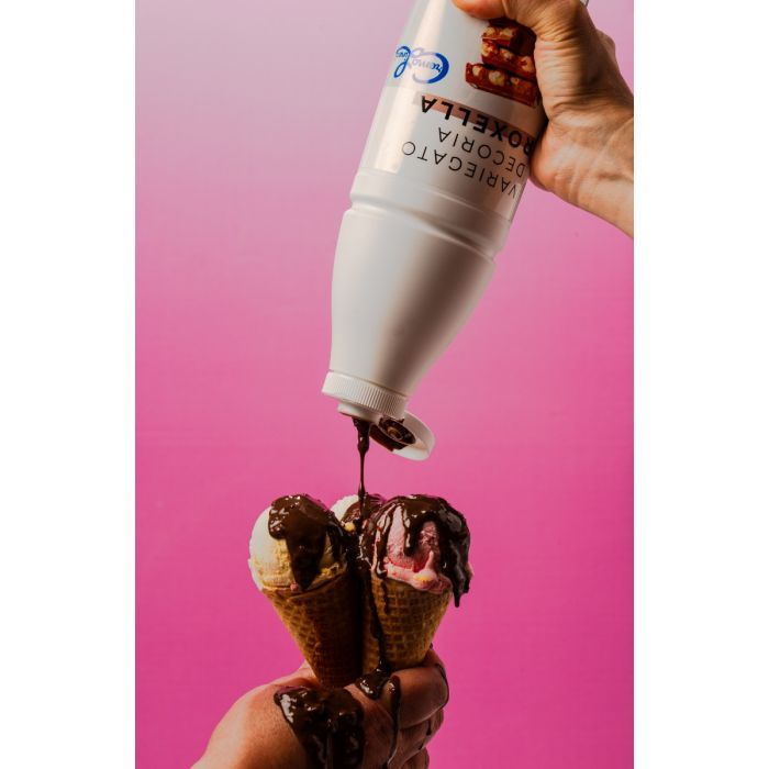 Chocolate syrup being poured from a white squeeze bottle onto two scoops of ice cream in waffle cones, with syrup dripping onto the hand.