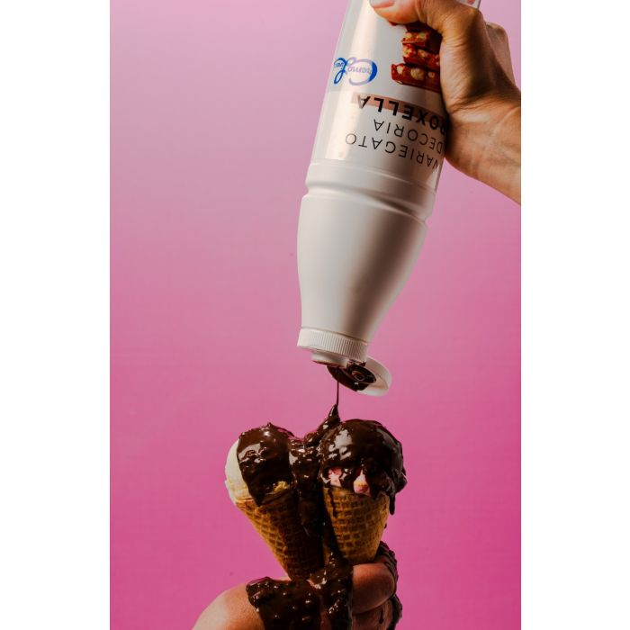 Hand pouring chocolate syrup from a white squeeze bottle onto two ice cream cones against a pink background.