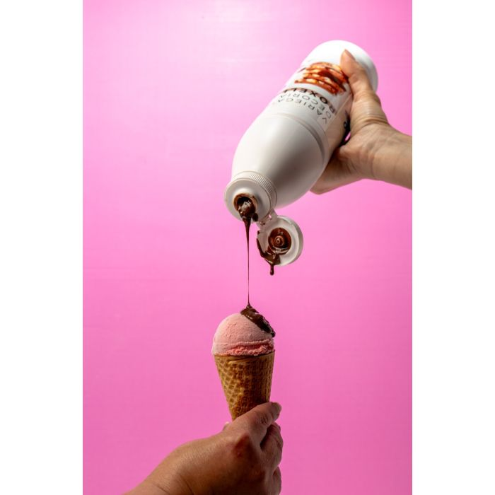 Chocolate syrup being poured from a white bottle onto a pink ice cream cone, with syrup dripping in a thin stream.