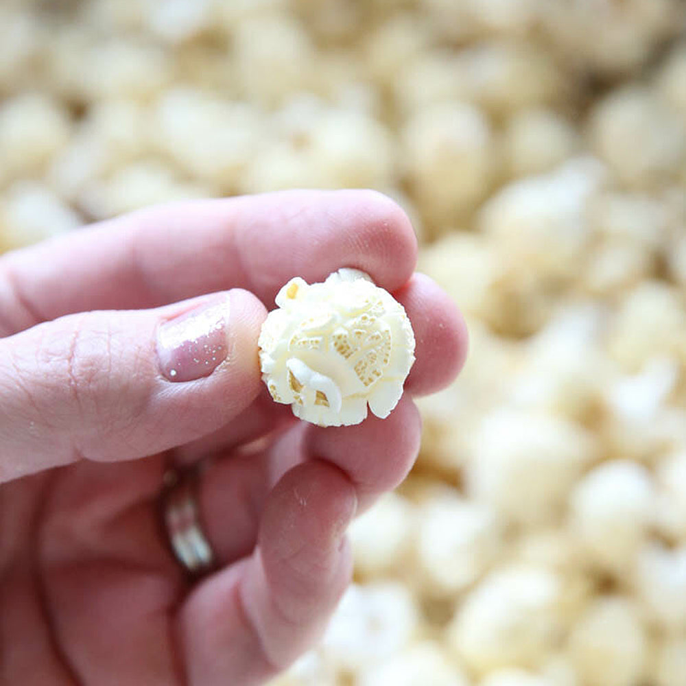 A person holding a piece of popcorn with many popcorn kernels in the background.