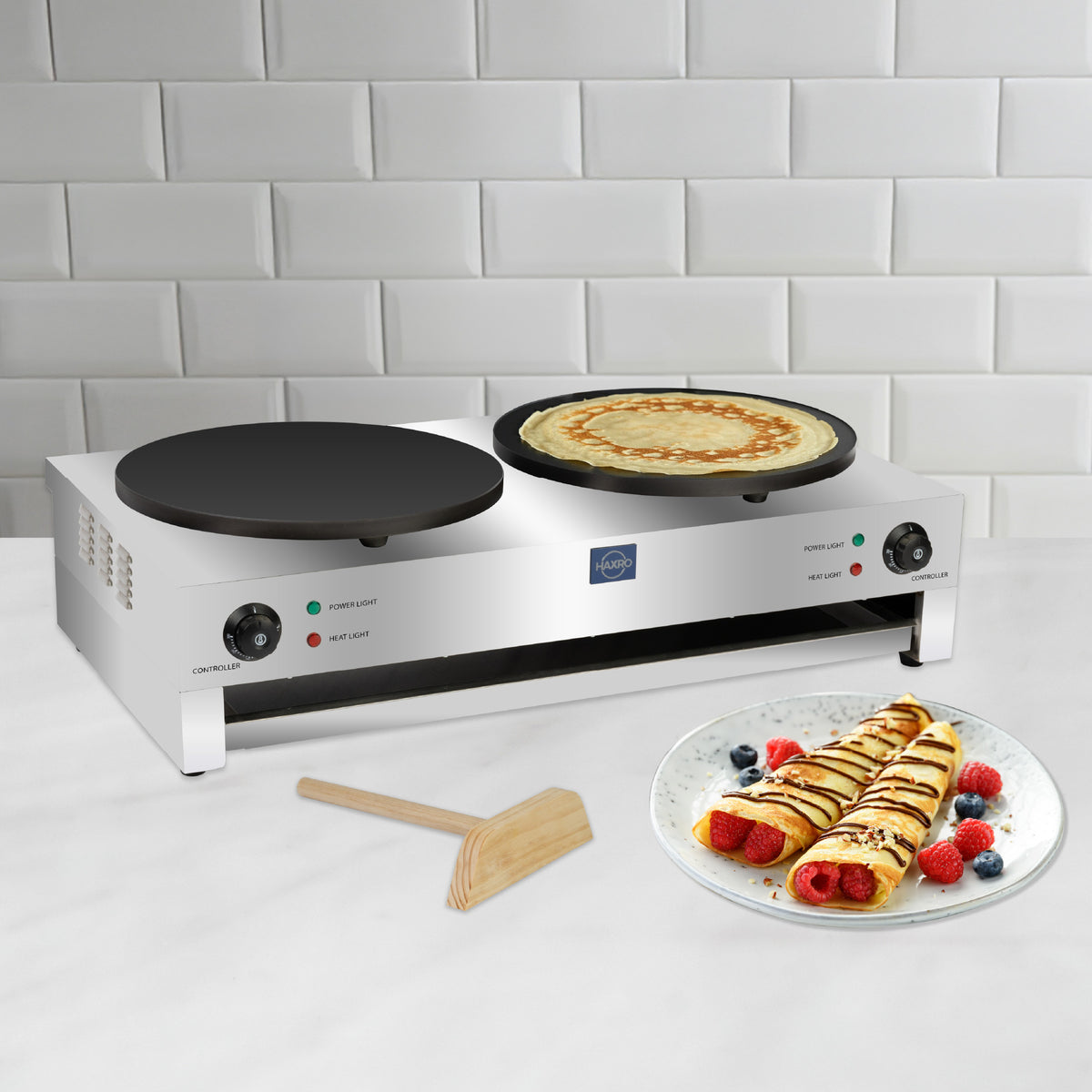 A sleek, double-burner crepe maker on a white countertop, with one crepe cooking. Nearby, a plate of rolled crepes topped with berries and chocolate drizzle.