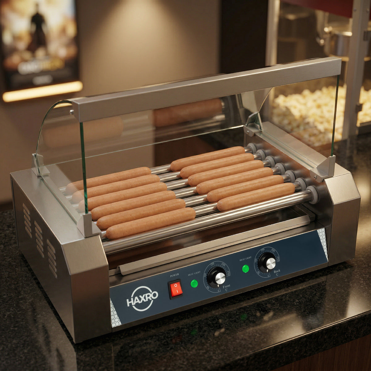 A stainless steel hot dog roller grill with several sausages cooking evenly. It has a glass cover and adjustable controls. Popcorn and a blurry movie poster are visible in the background, suggesting a snack area or cinema setting.