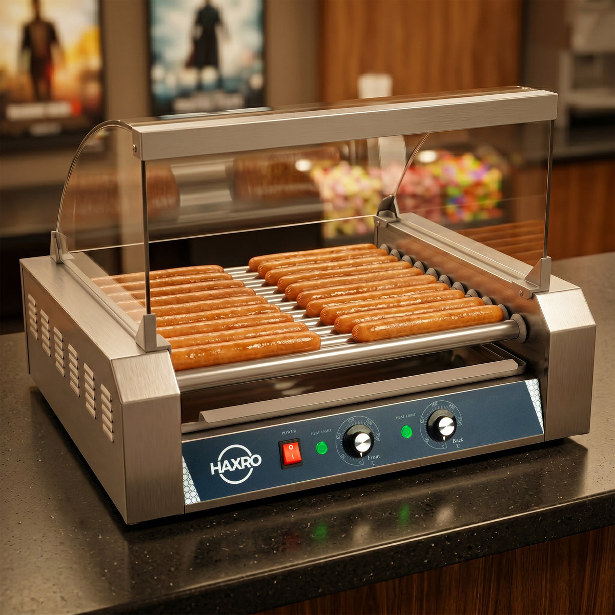 A hot dog roller grill with about 20 hot dogs evenly spaced on rotating bars. The stainless steel machine has controls on the front panel and is set on a black countertop. The background shows blurred movie posters, creating a casual concession stand vibe.