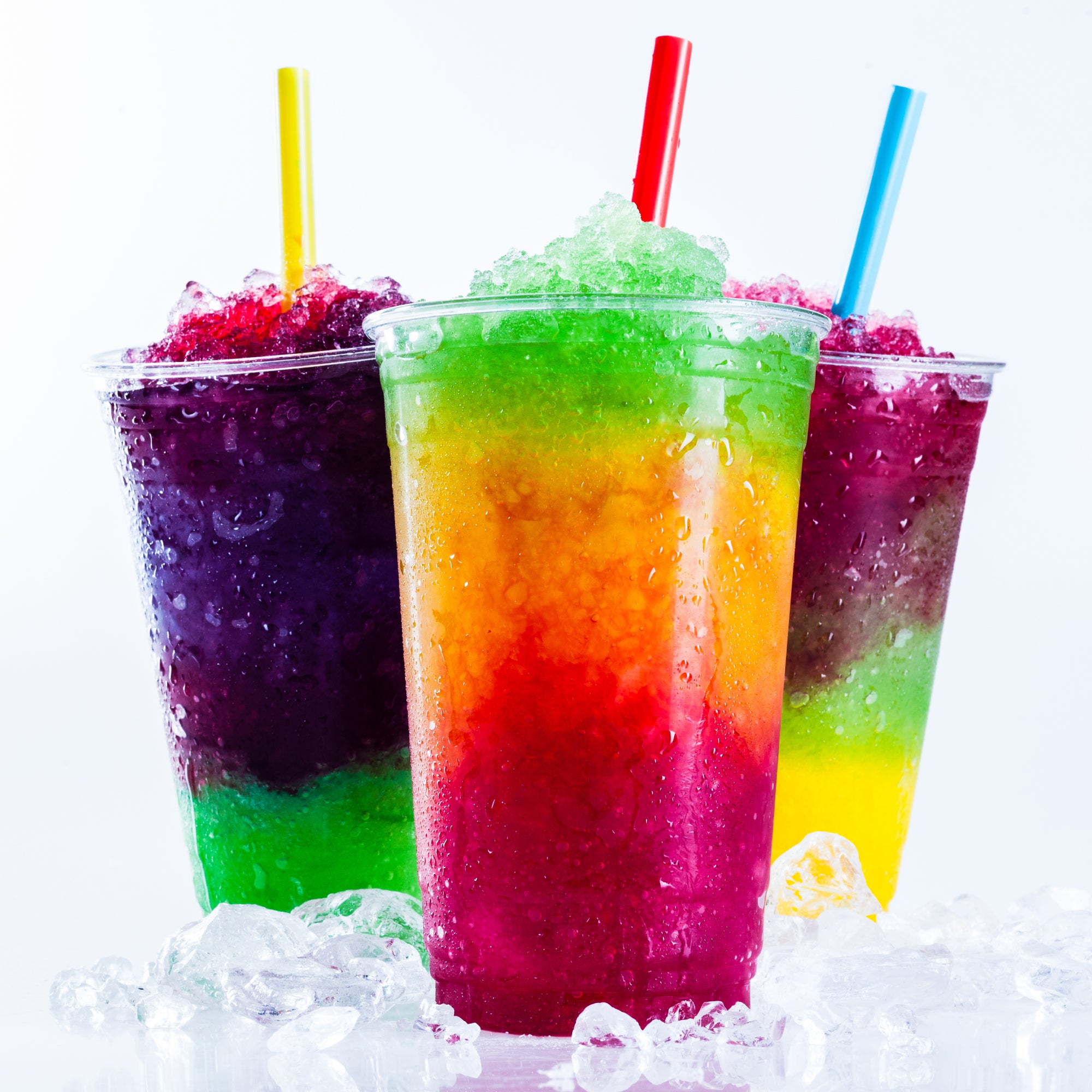 Three colorful slushies in clear cups with straws. The vibrant layers include purple, green, orange, and red, surrounded by ice on a white background. Refreshing and playful.
