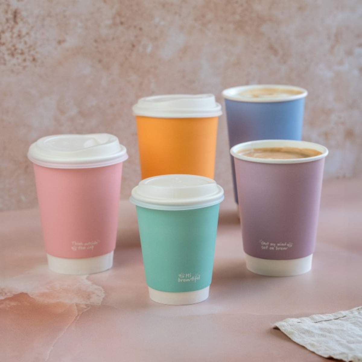 Five colorful takeaway coffee cups in various pastel shades arranged on a pink surface.