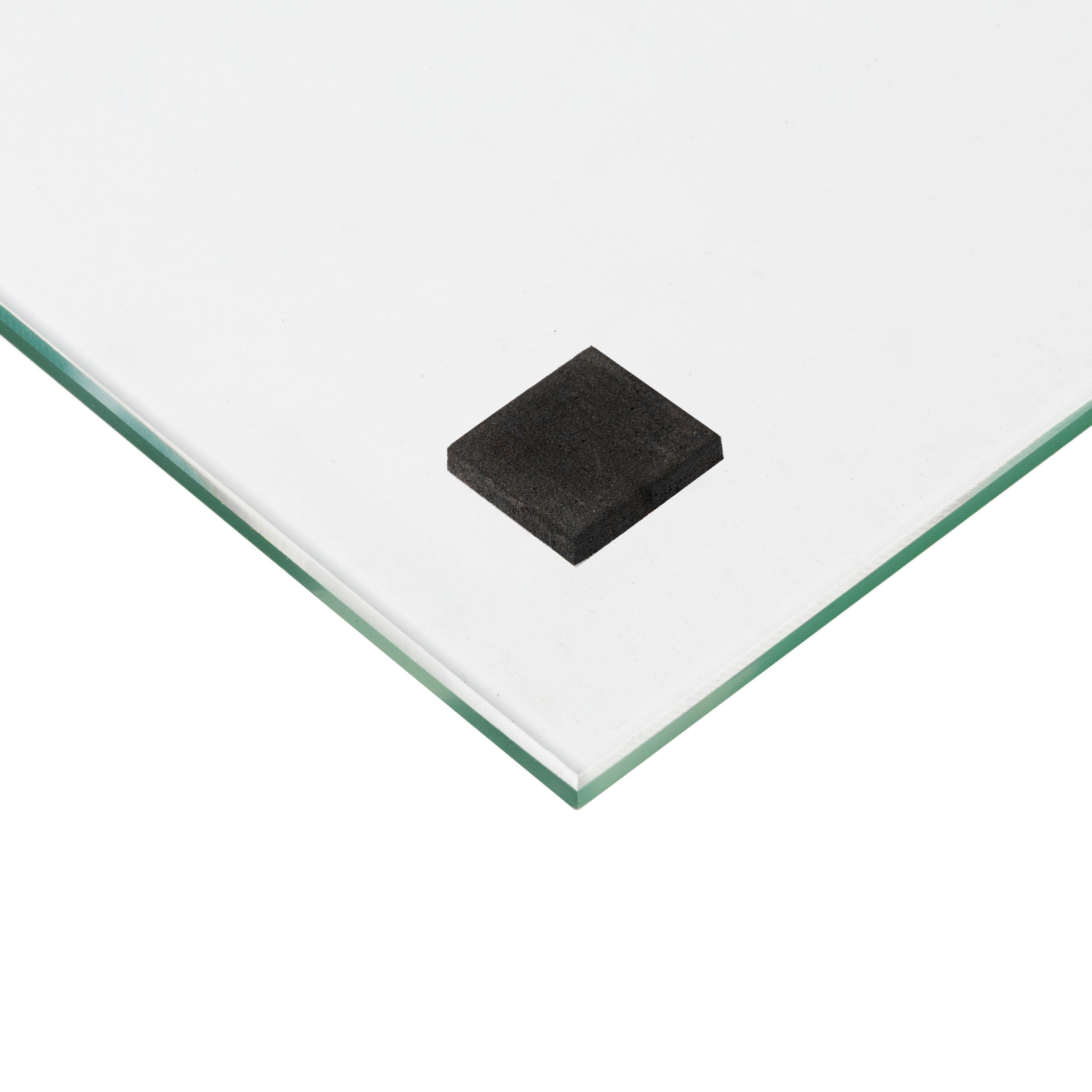 Close-up of a glass corner with a small black square pad attached underneath. The glass has smooth edges and rests against a white background.