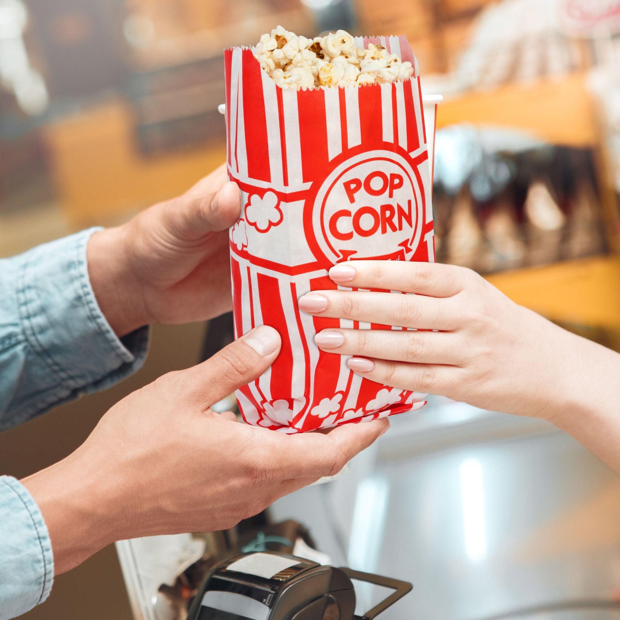 Two hands exchange a red and white striped popcorn bag in a warm, cheerful setting. The bag is filled with popcorn, highlighting a cinema or festive atmosphere.