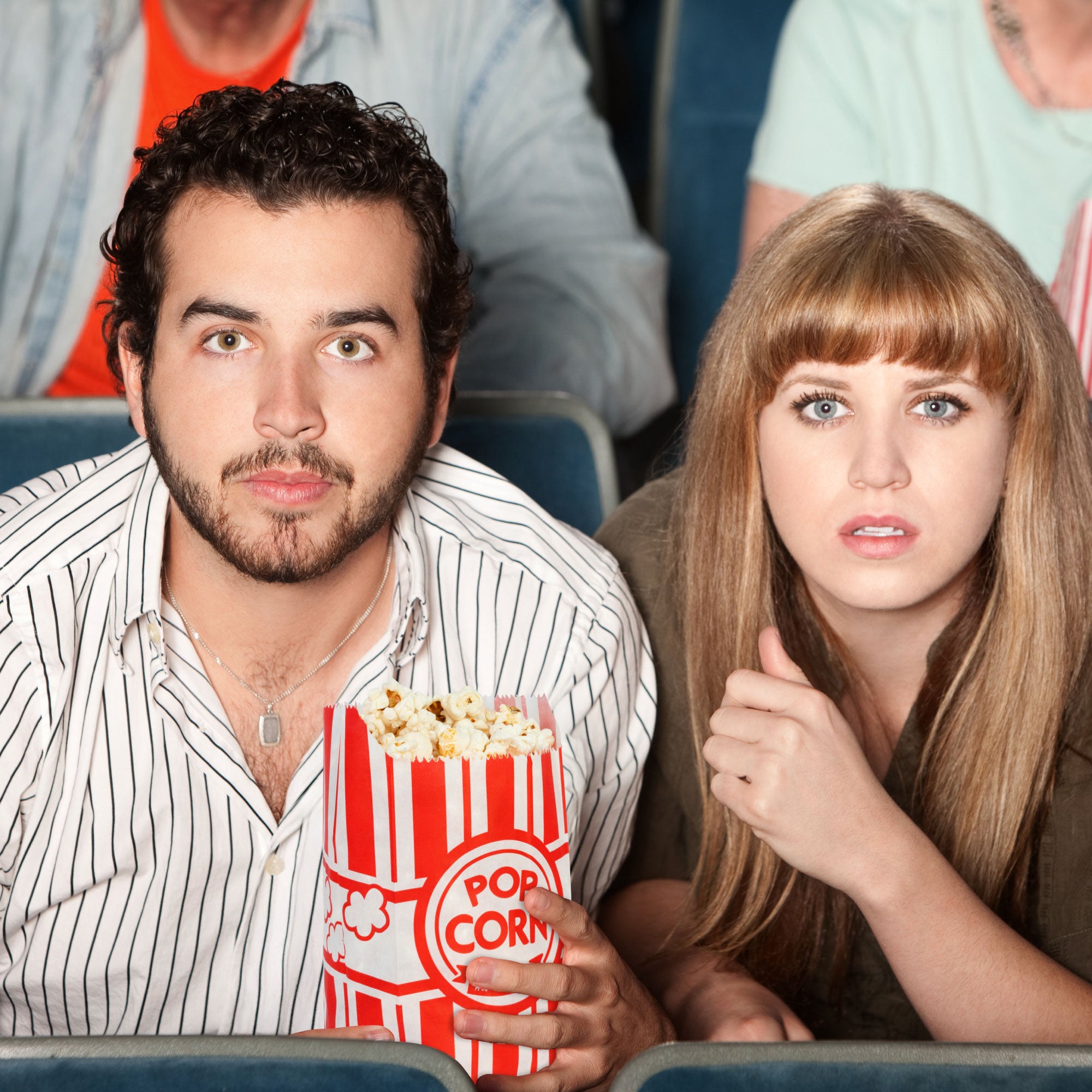 A man and woman sit in a theater, wide-eyed and focused. The man holds popcorn. Their expressions suggest intense interest or surprise.