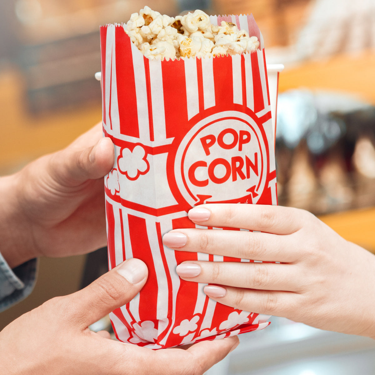 A hand holding a red and white striped popcorn bag filled with popcorn, being offered to another hand.