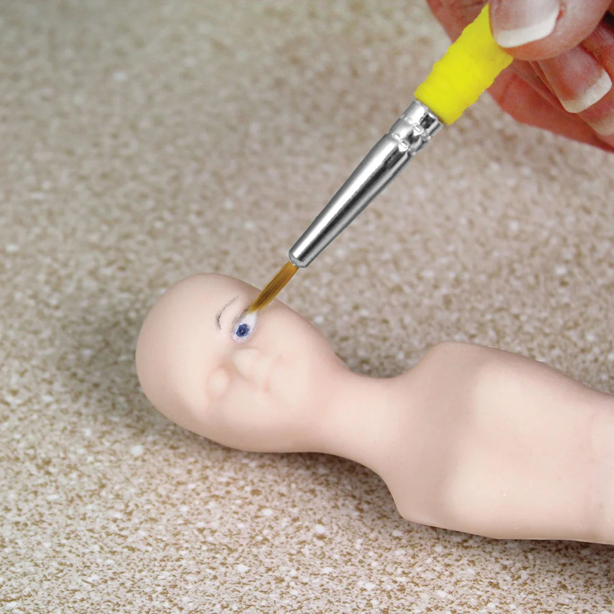 A close-up of a hand using a fine paintbrush with a yellow handle to apply blue and white eye makeup to a small beige doll head, set against a textured beige carpet background.