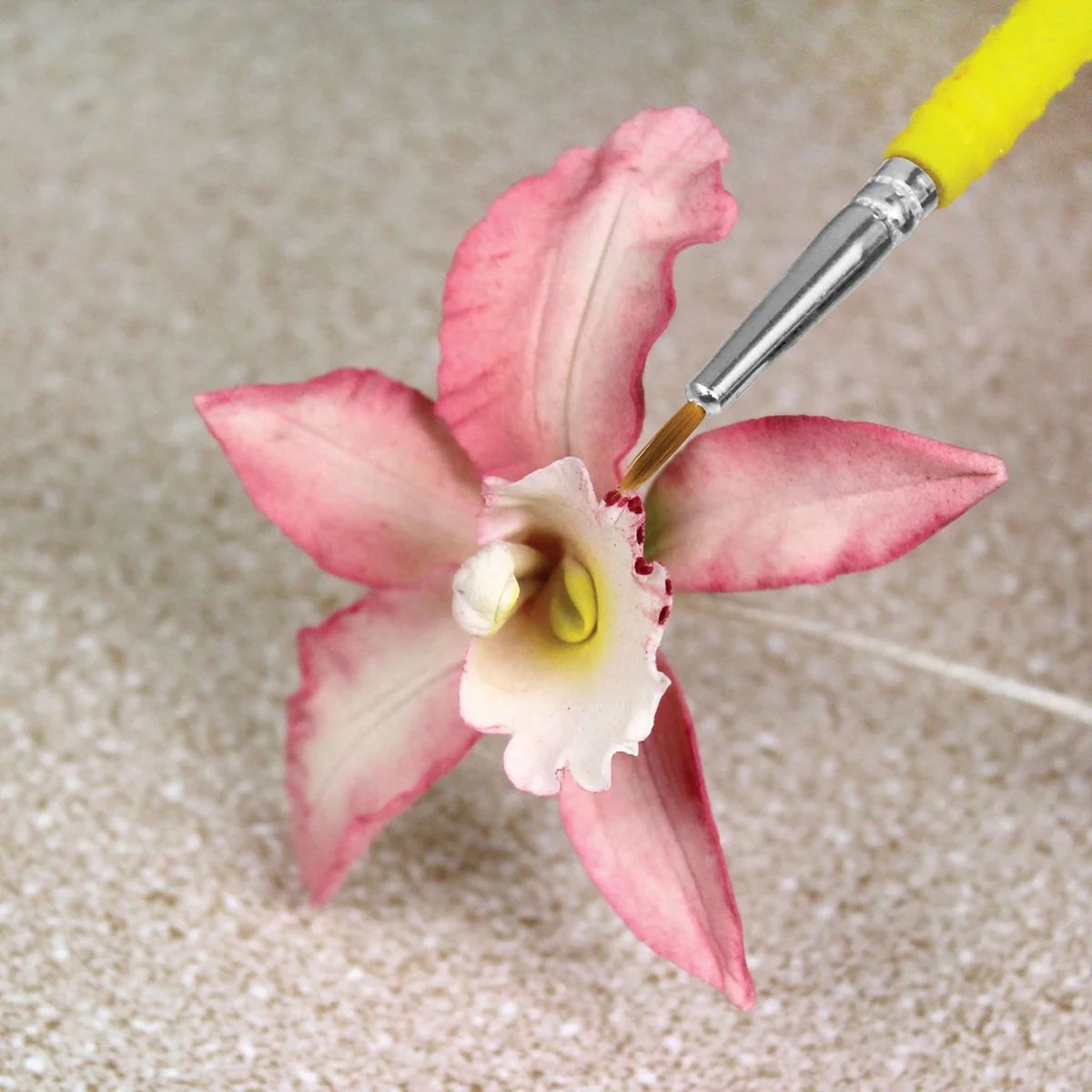 A pink and white artificial orchid flower with a yellow center, shown close-up on a beige textured surface with a small yellow-handled paintbrush touching the center.