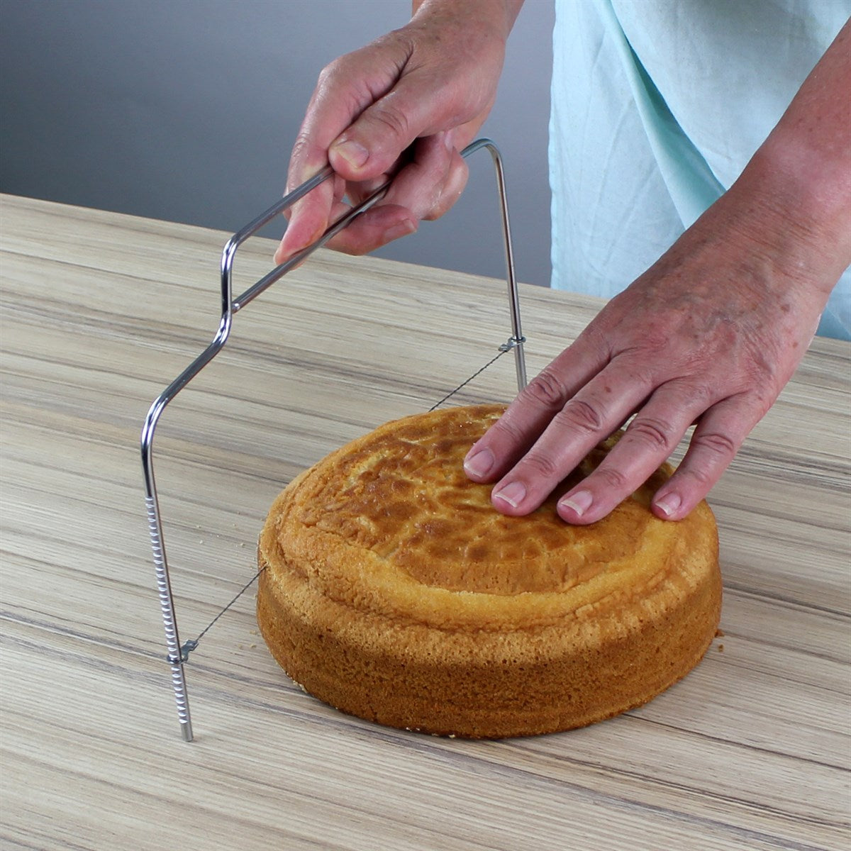 A person slices a round sponge cake with a metal cake leveler on a wooden surface. Hands are steady, conveying precision and focus.