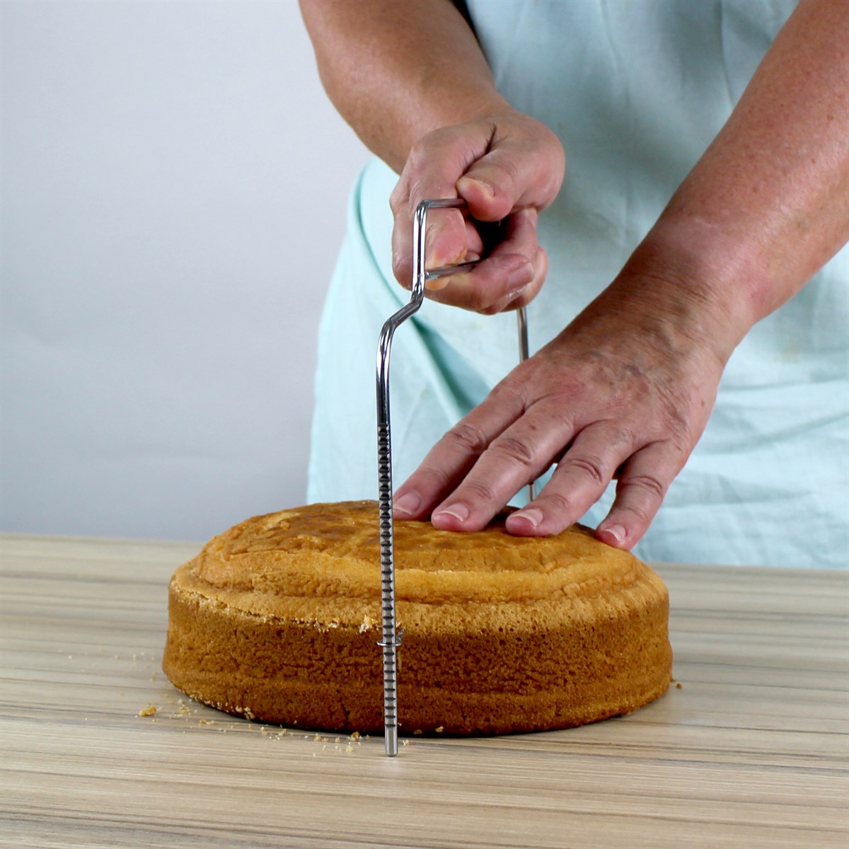 A person in a light blue apron uses a cake leveler to slice a round cake on a wooden table, focusing intently on creating an even layer.