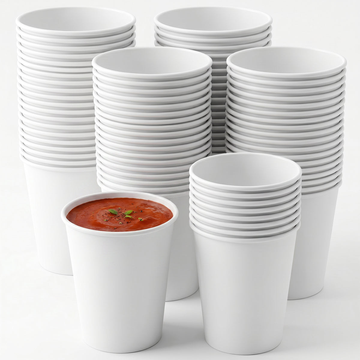 A stack of white paper cups surrounds a single cup filled with tomato soup, garnished with herbs. The scene is minimalist and orderly.