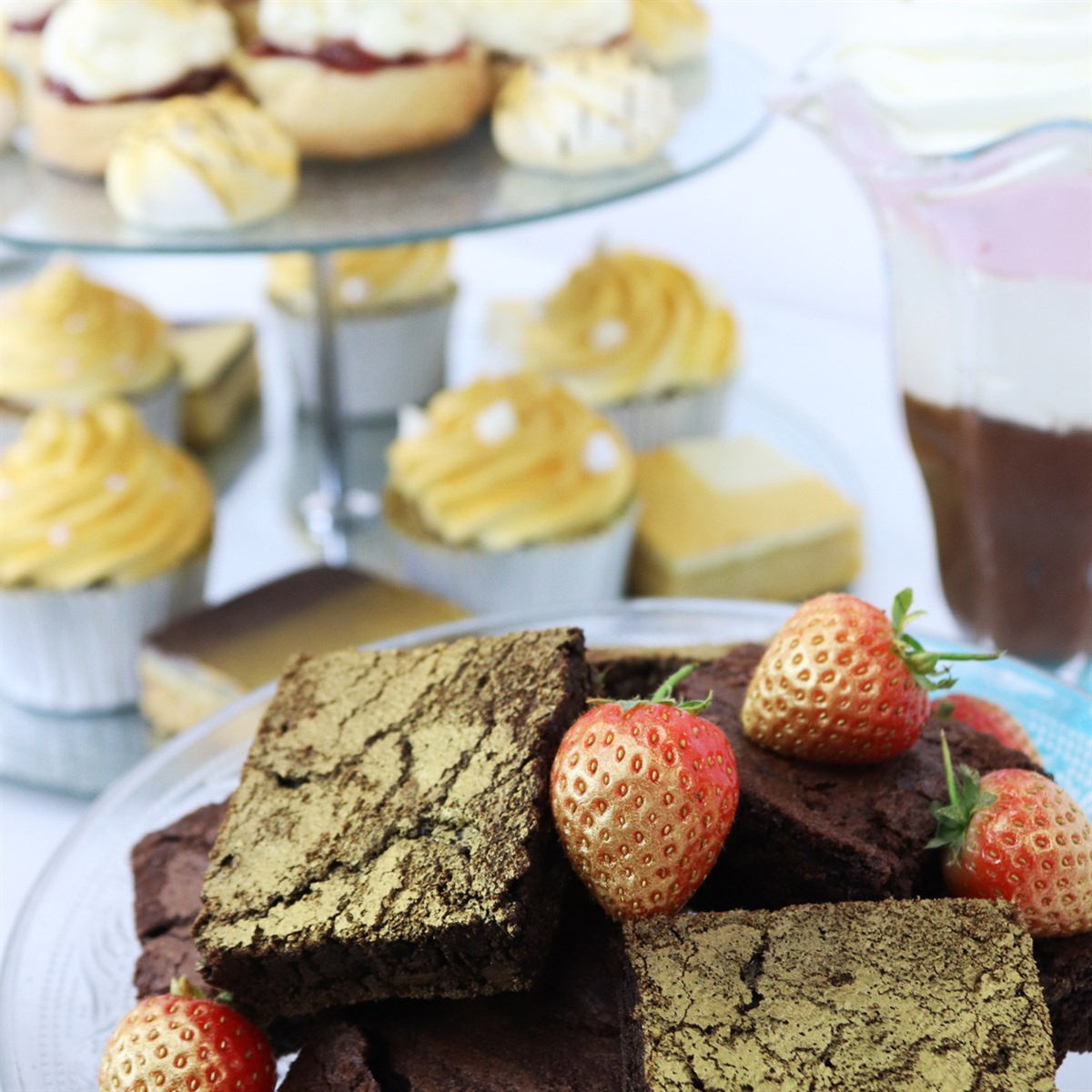 A dessert table featuring gold-dusted brownies topped with strawberries, cupcakes, and layered pastries. The scene conveys a sense of indulgence and celebration.
