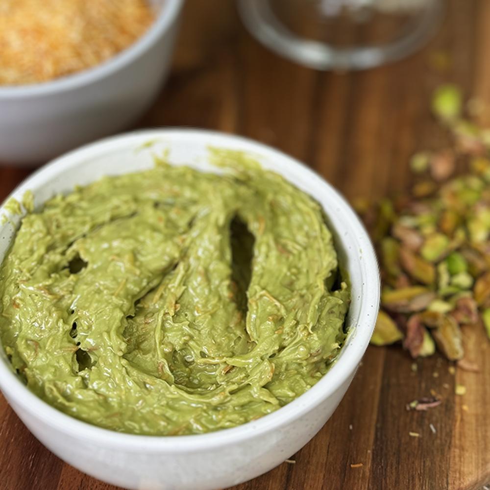 A white bowl filled with creamy, green pistachio butter on a wooden surface. Crushed pistachios are scattered nearby, adding a rustic feel.