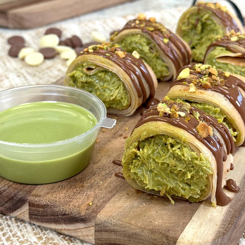 Slices of pistachio-filled dessert rolls drizzled with chocolate, topped with nuts, are displayed on a wooden board. A small cup of green sauce is on the side.