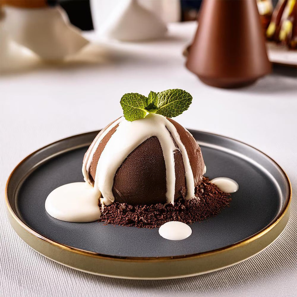 A chocolate dome dessert drizzled with white chocolate sauce sits on a black plate, garnished with fresh mint. The setting is elegant and inviting.