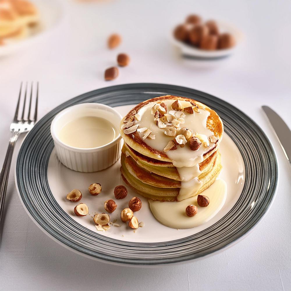 A stack of pancakes with cream sauce and chopped nuts sits on a plate. A small bowl of extra sauce is beside them. The setting is bright and appetizing.