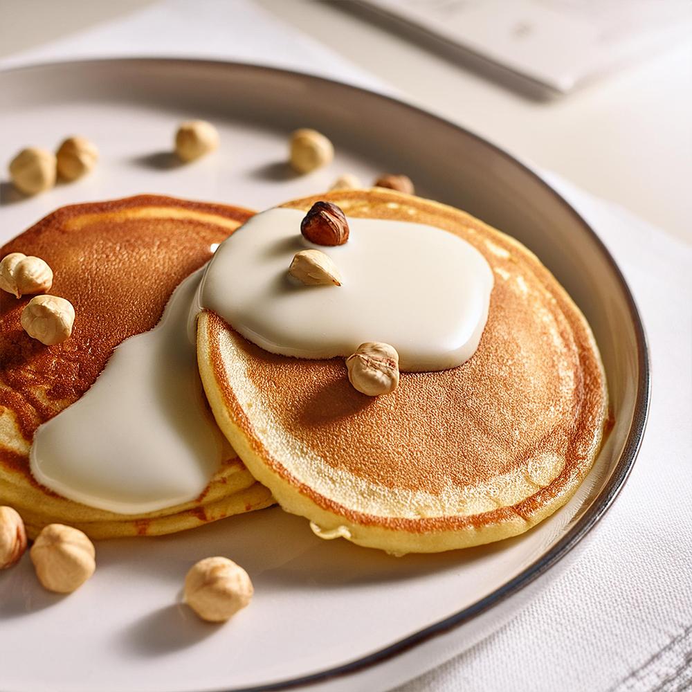 Two golden pancakes topped with smooth cream and hazelnuts on a white plate, creating a warm and inviting breakfast scene.