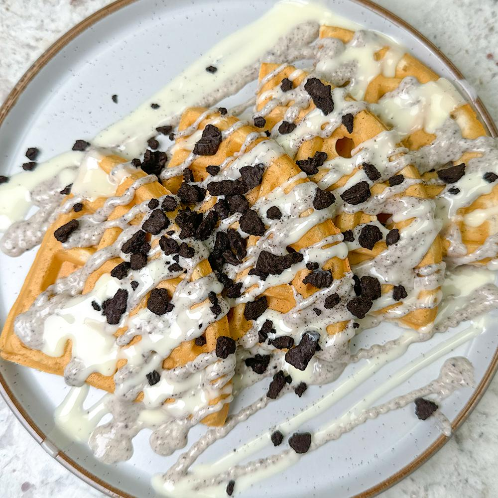 Plate of golden waffles topped with white and cookies-and-cream drizzle, sprinkled with crushed chocolate cookie pieces.