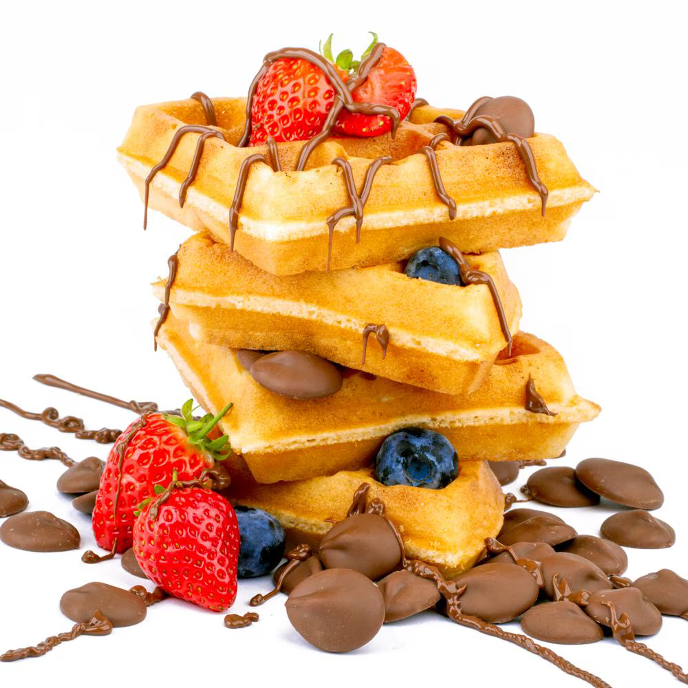A stack of waffles topped with fresh strawberries, blueberries, and drizzled with chocolate syrup, surrounded by chocolate-coated candy.