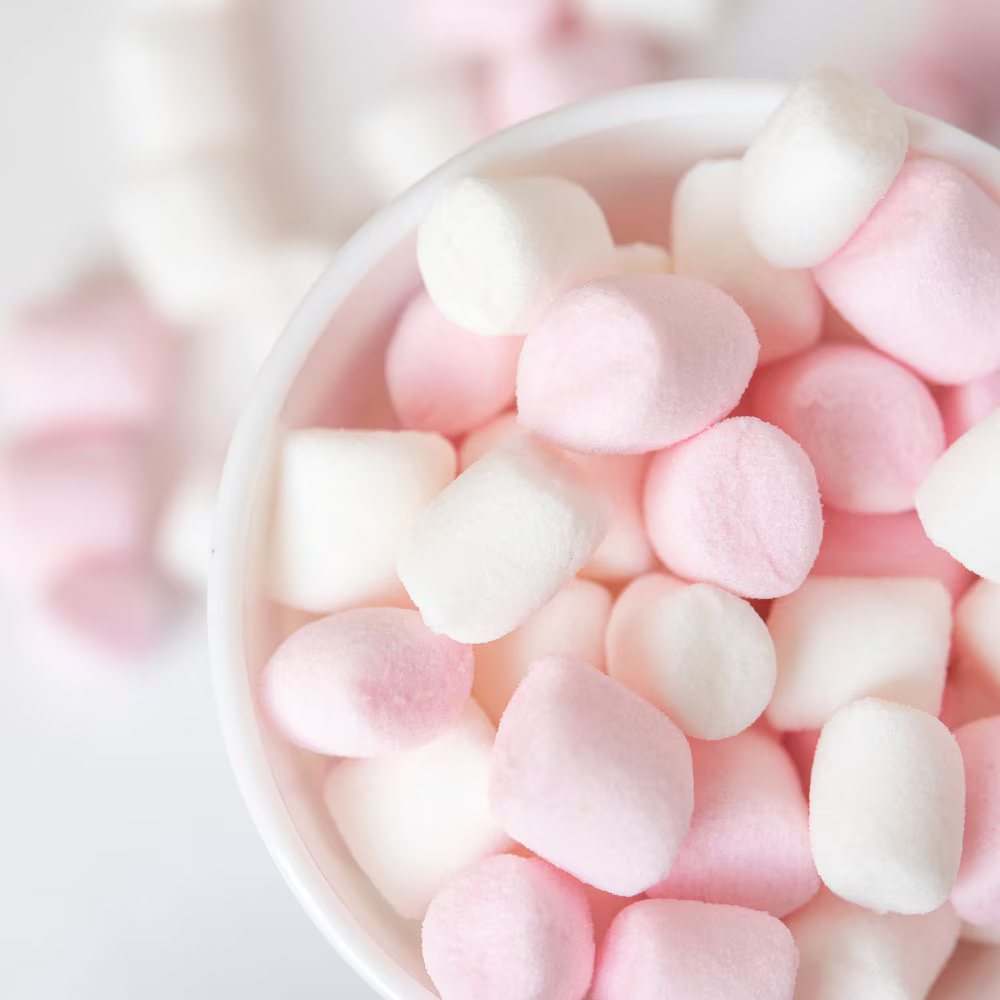 A close-up view of a bowl filled with pink and white mini marshmallows, with more marshmallows softly blurred in the background.