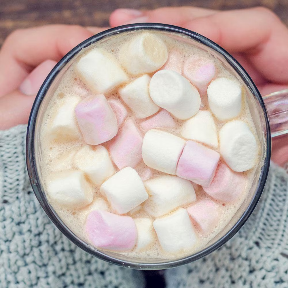 The image shows a close-up view of a warm cup of hot chocolate topped with fluffy white and pink marshmallows. The cup is held by hands wearing a cozy knitted sweater. The marshmallows are scattered generously on the surface of the drink, adding a soft, sweet touch to the beverage.