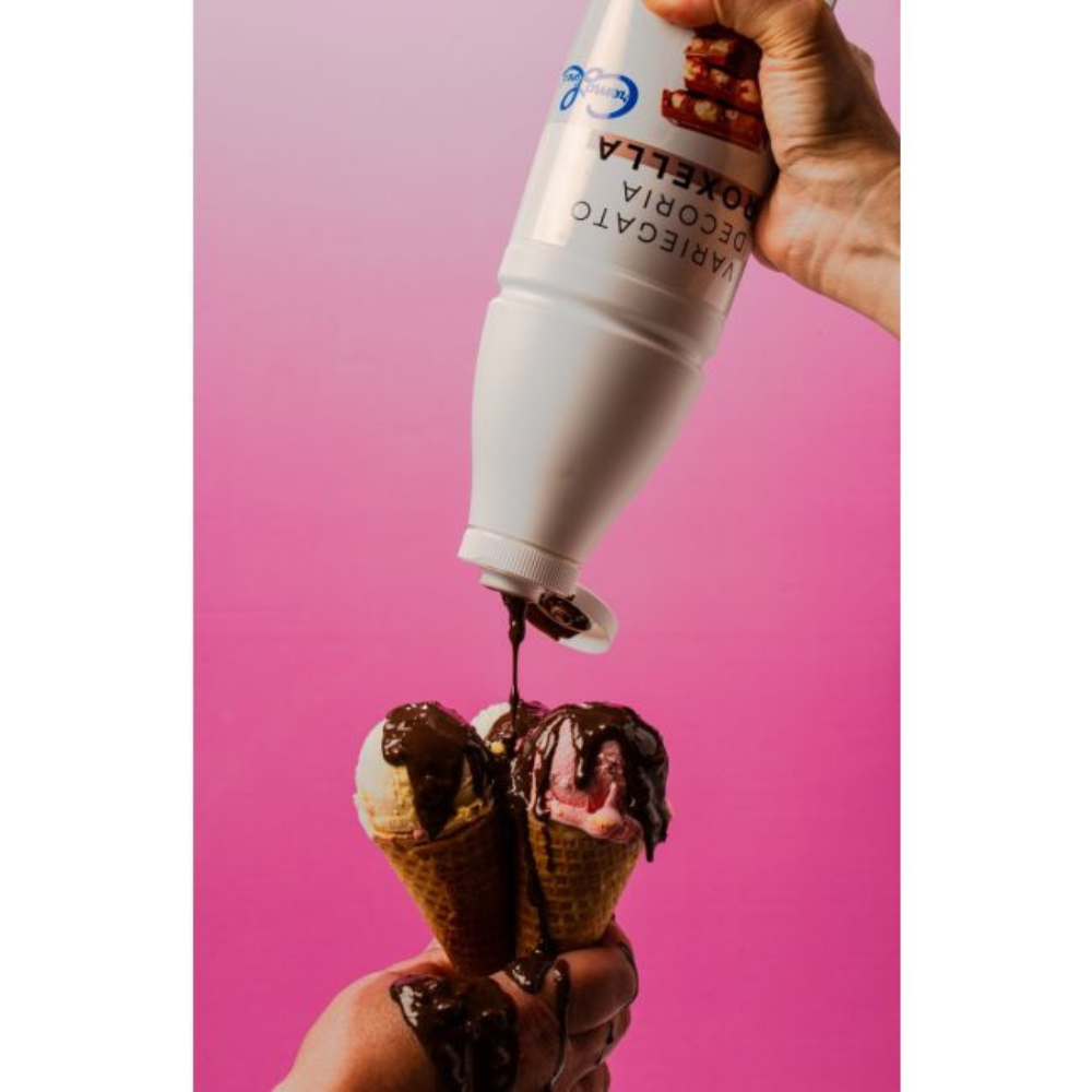 A hand pours chocolate syrup onto three ice cream cones held together against a vibrant pink background, creating a playful, indulgent scene.
