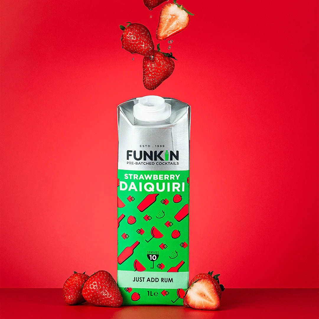 A carton of Funkin Strawberry Daiquiri cocktail mixer stands against a bright red background, with fresh strawberries falling above it and several strawberries placed at its base.
