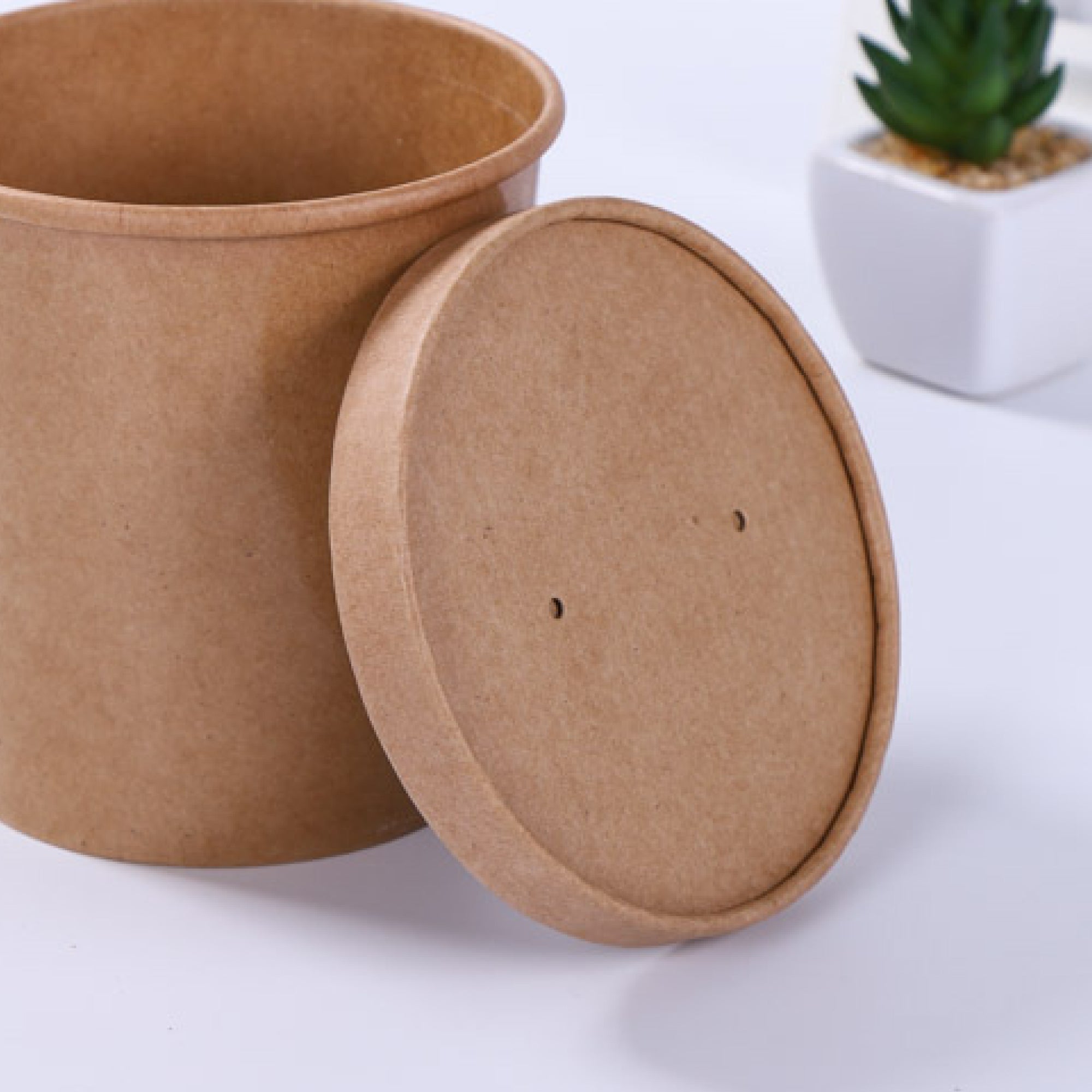 A brown kraft paper container with a removable lid, placed on a flat surface beside a small potted plant.