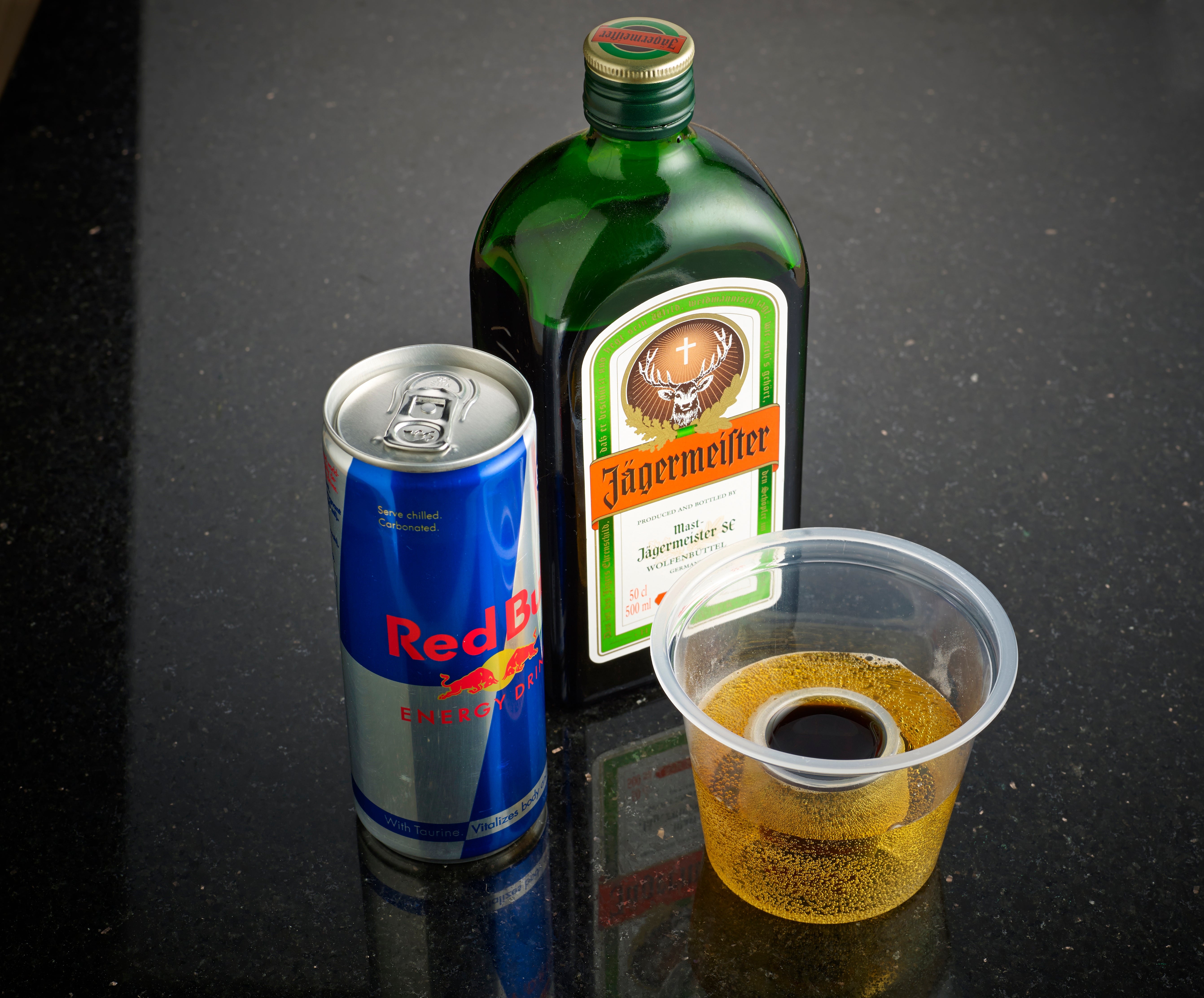 A can of Red Bull, a bottle of Jägermeister, and a clear plastic cup filled with a brown liquid on a black surface.