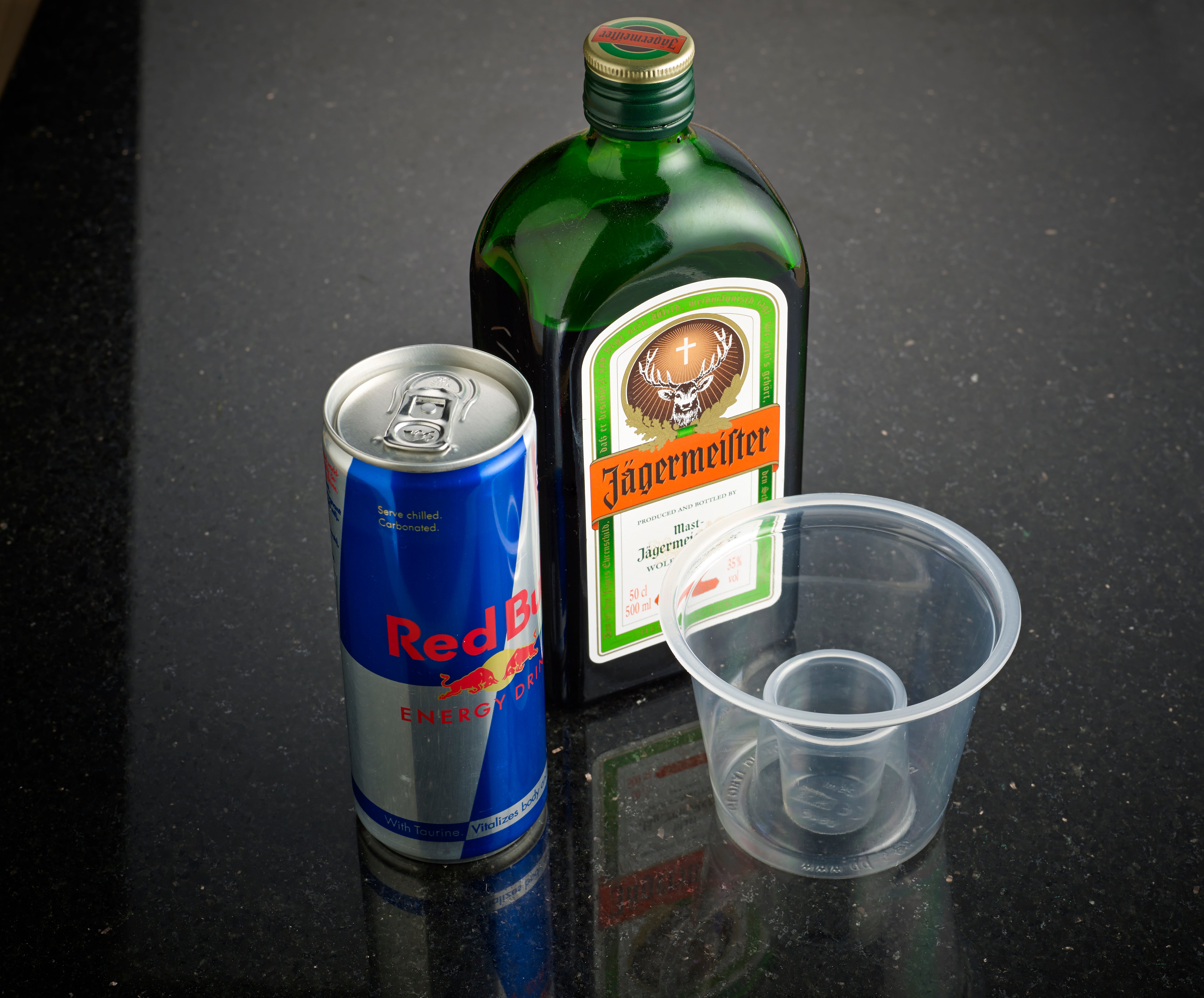 A can of Red Bull, a bottle of Jägermeister, and a plastic cup on a black surface.