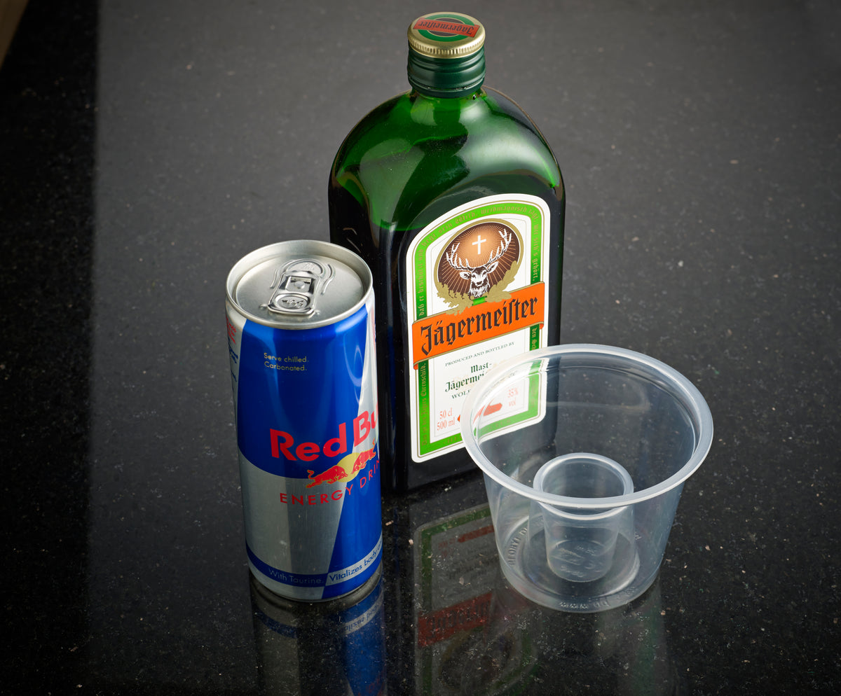 A can of Red Bull, a bottle of Jägermeister, and a plastic cup on a black surface.