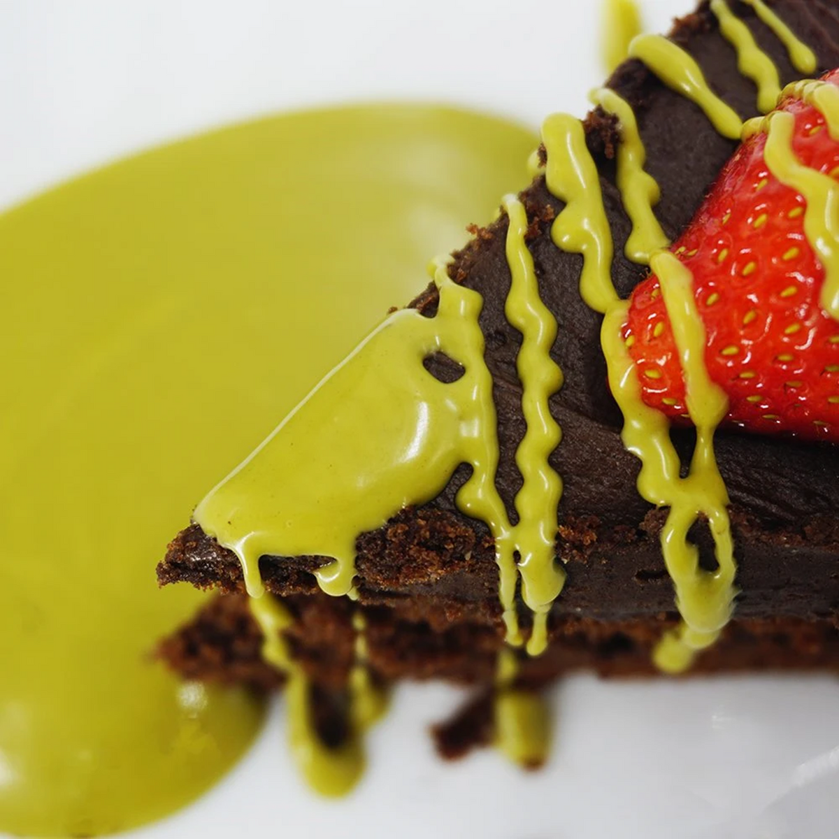 A close-up of a chocolate cake slice drizzled with green matcha glaze, topped with fresh strawberries. The cake rests on a white plate.