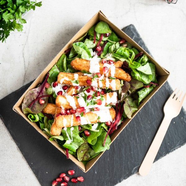 Salad with greens, pomegranate seeds, and croutons in a cardboard container on a slate surface.