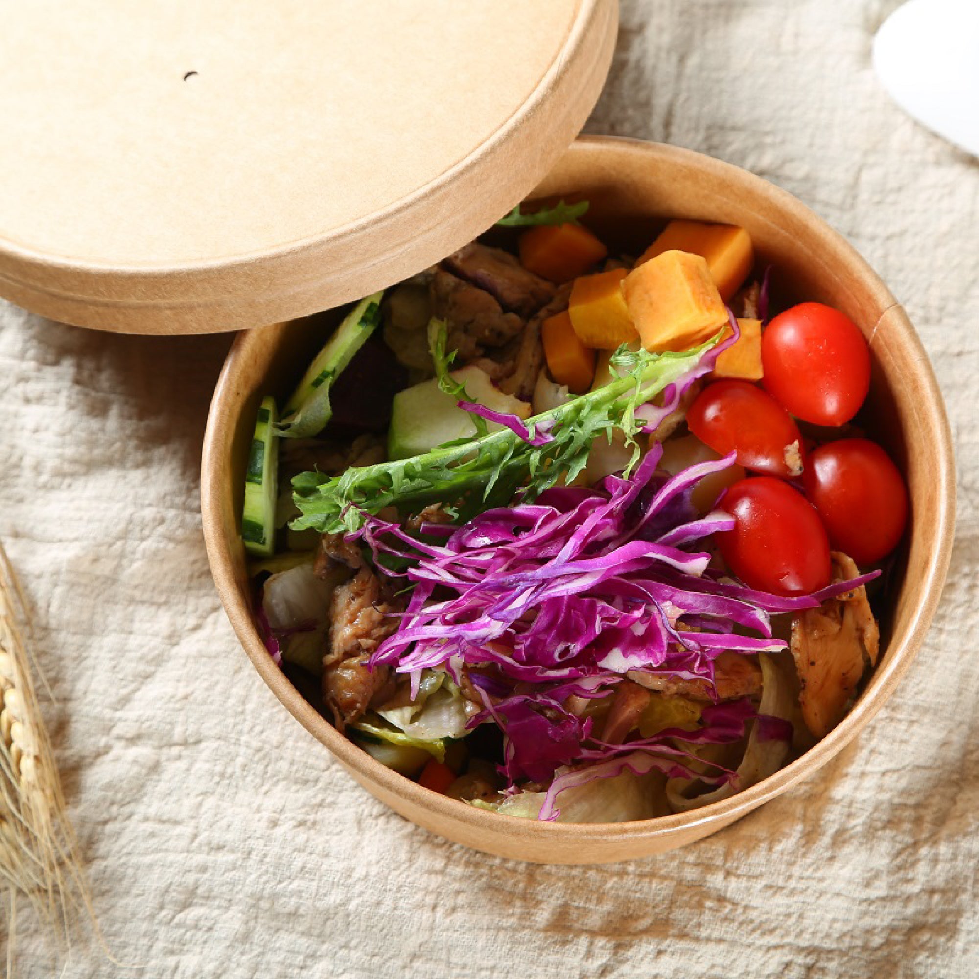 A round Kraft bowl with its lid off, filled with a colorful salad including red cabbage, tomatoes, cucumbers, and sweet potato on a textured beige fabric background.