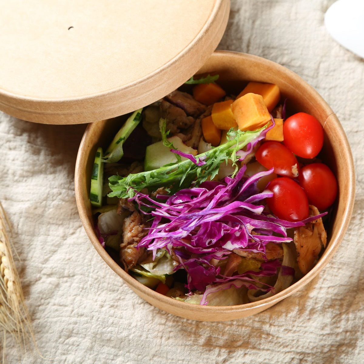 A round Kraft bowl with its lid off, filled with a colorful salad including red cabbage, tomatoes, cucumbers, and sweet potato on a textured beige fabric background.