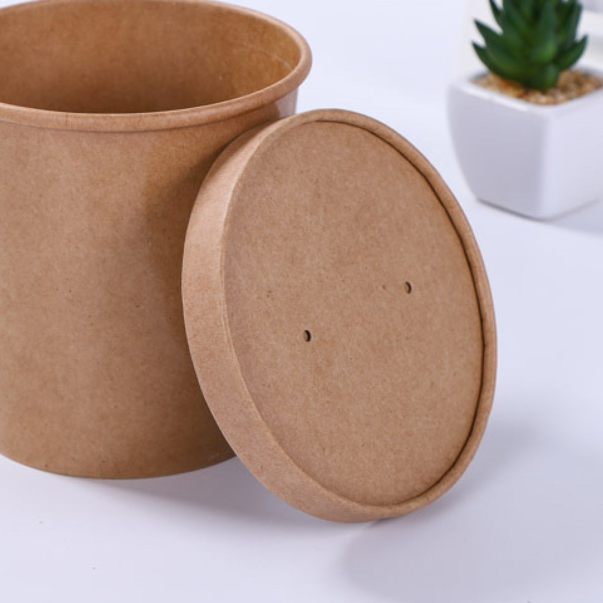 A brown kraft paper container with a removable lid, placed on a flat surface beside a small potted plant.