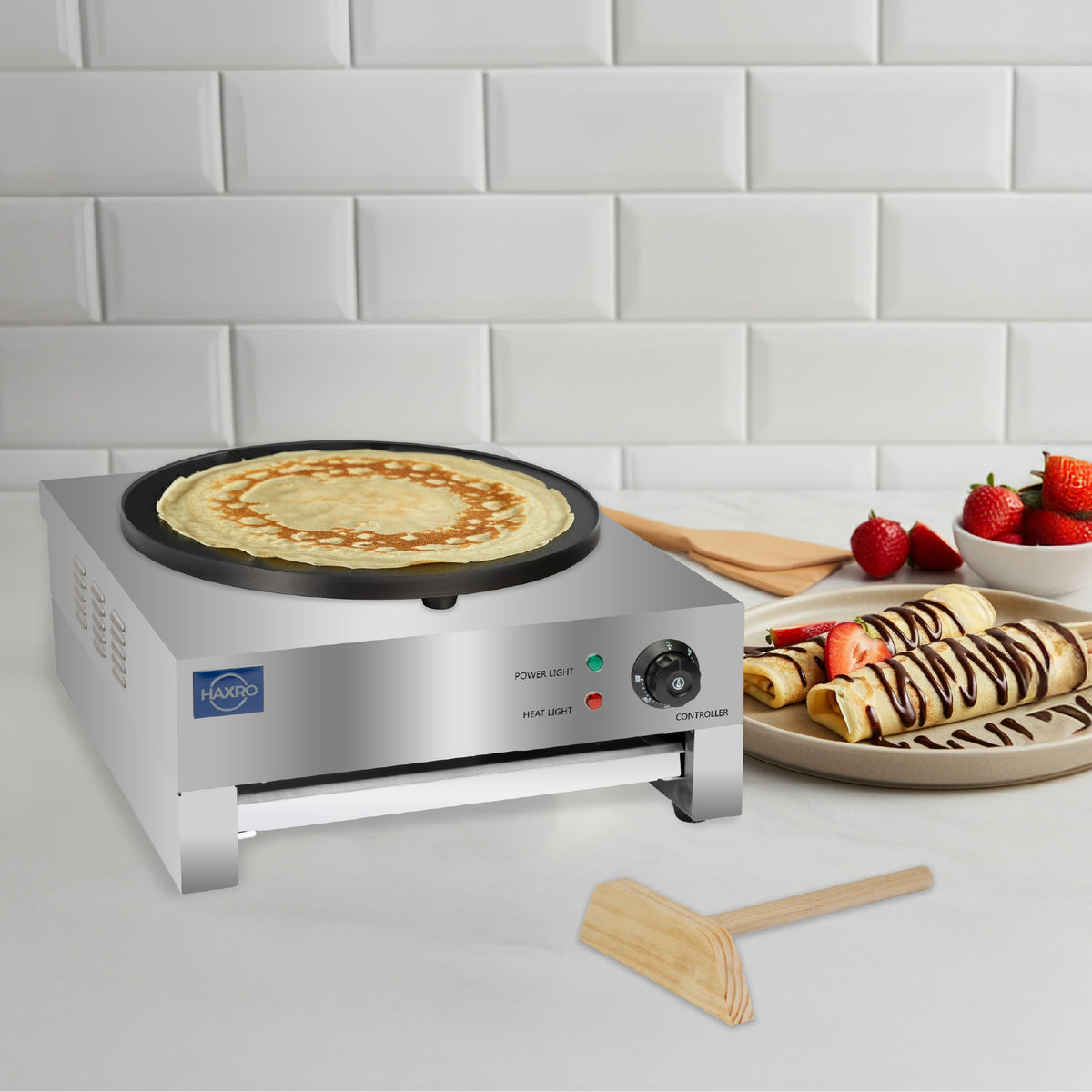 Haxro crepe maker cooking a crepe on a kitchen counter, with strawberries and chocolate-drizzled rolled crepes displayed on a plate beside it.