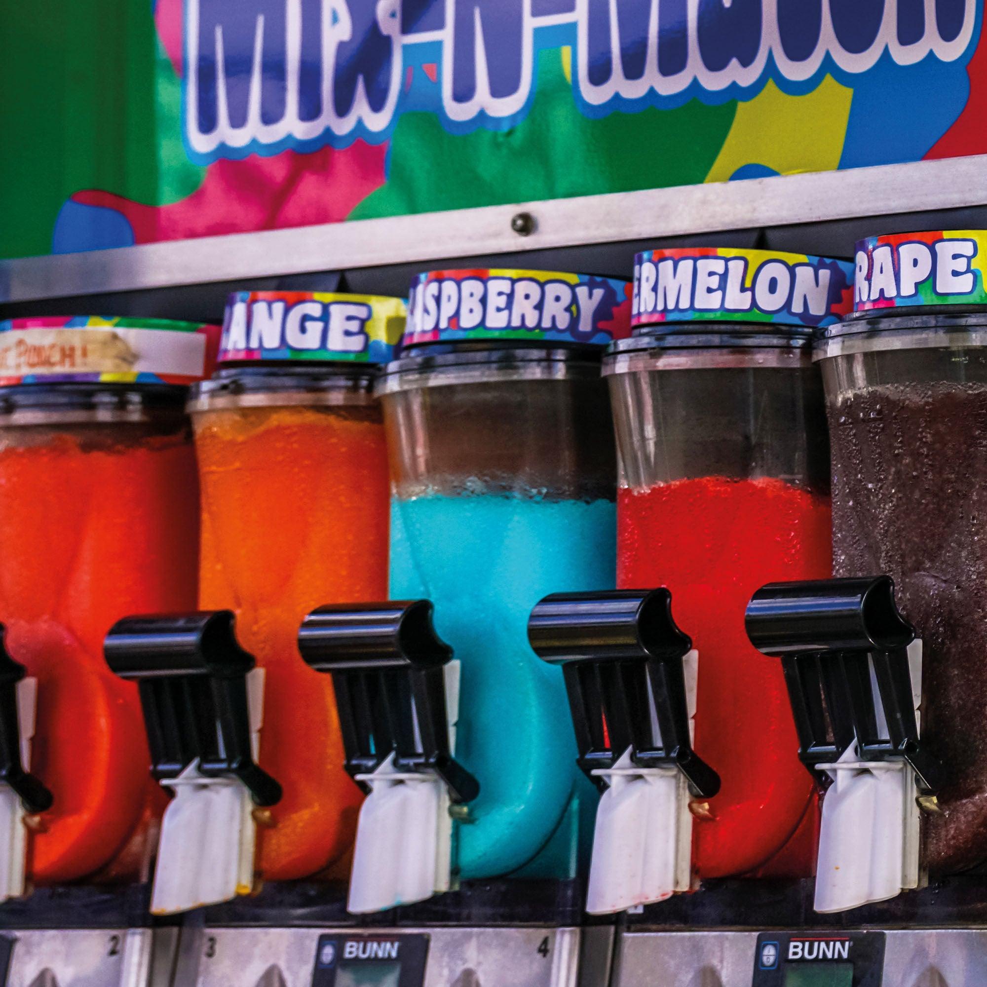 A close-up shot of several slush machine dispensers filled with brightly colored slush flavors, including orange, blue, red, and purple. Each dispenser has a black nozzle labeled with a white tag, and colorful labels with flavor names like "Orange," "Raspberry," "Watermelon," and "Grape."