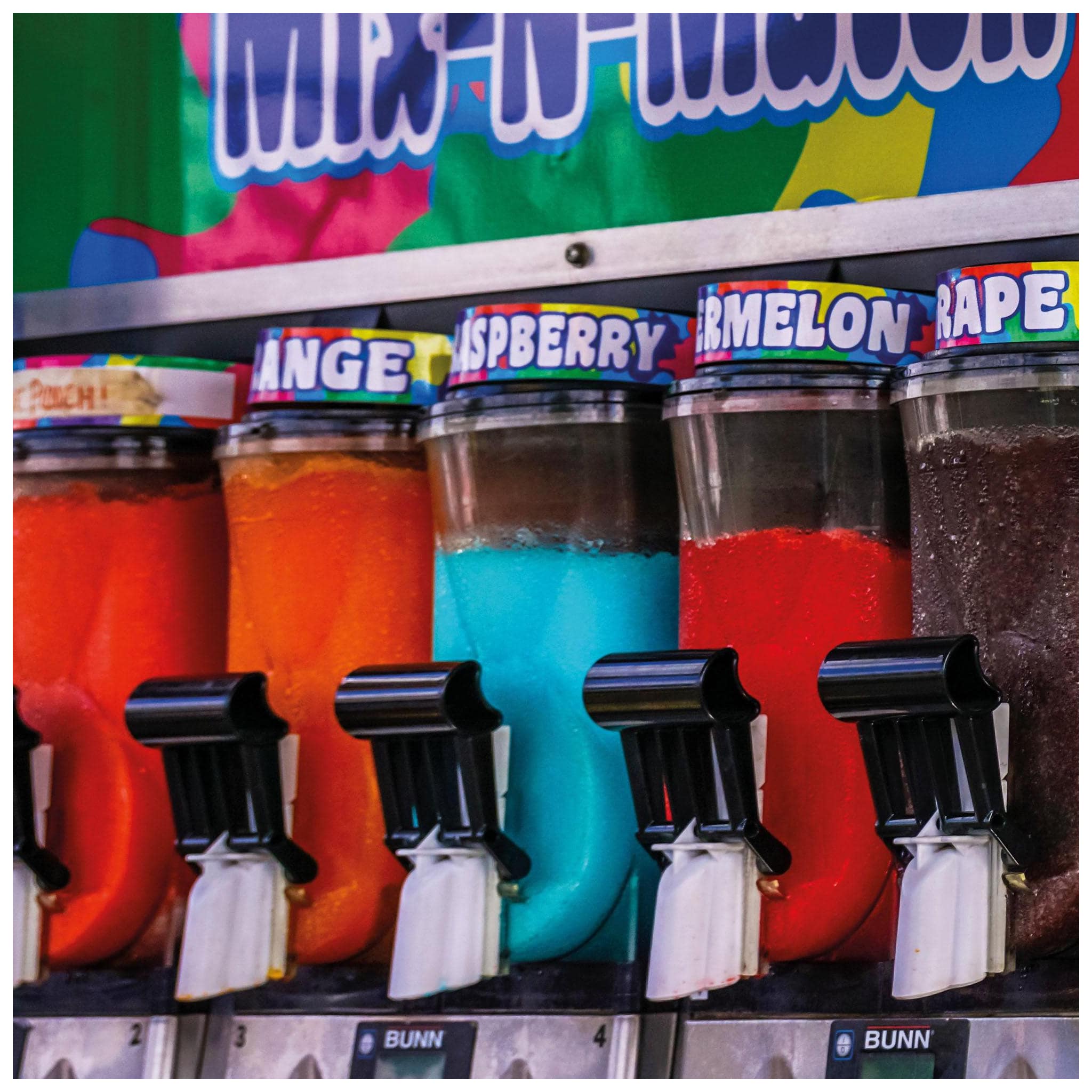 A close-up shot of several slush machine dispensers filled with brightly colored slush flavors, including orange, blue, red, and purple. Each dispenser has a black nozzle labeled with a white tag, and colorful labels with flavor names like "Orange," "Raspberry," "Watermelon," and "Grape."