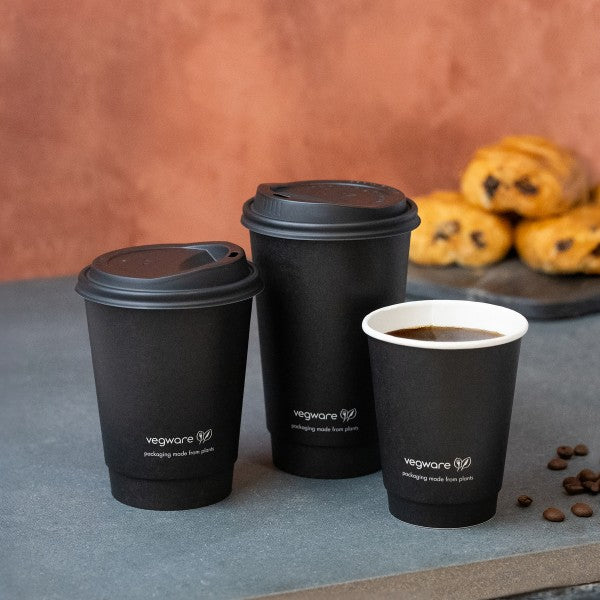A collection of three black disposable coffee cups of varying sizes, with the smallest cup being white on the inside, placed on a gray surface. In the background, there are baked goods and coffee beans.