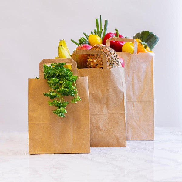 Three brown paper bags filled with various fruits and vegetables, including herbs, sitting on a marble surface.