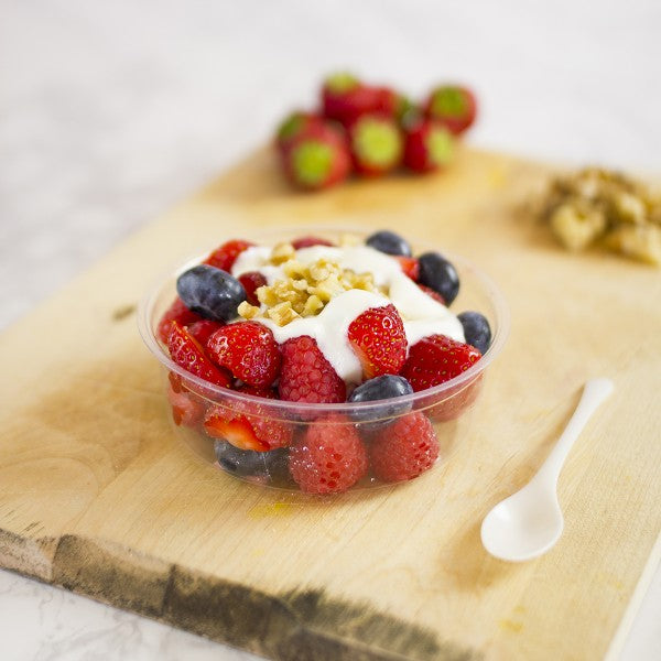 A plastic cup filled with mixed berries, yogurt, and nuts, placed on a wooden cutting board with a small spoon beside it.