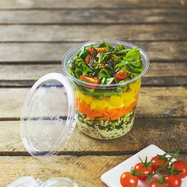 A clear cup filled with a layered salad consisting of greens, chopped vegetables, and cherry tomatoes, placed on a wooden surface.