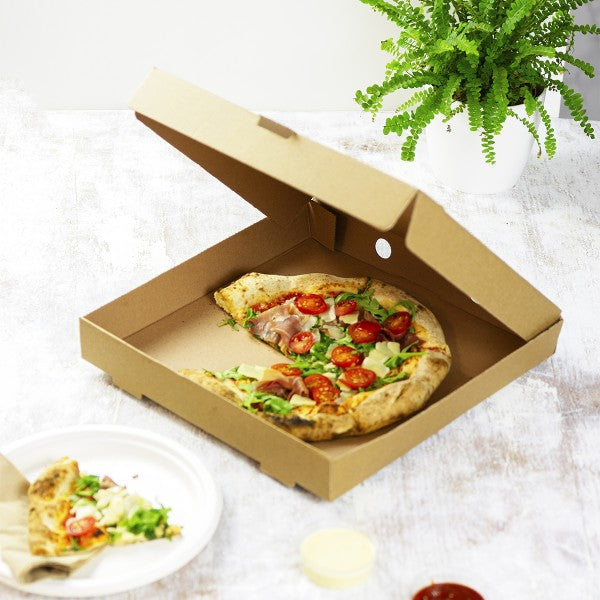 Open cardboard pizza box with a pizza inside, on a light background with a plant.