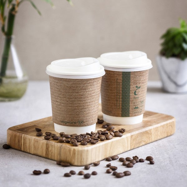 Two coffee cups with paper sleeves on a wooden serving board, surrounded by coffee beans, with a blurred background featuring a plant.