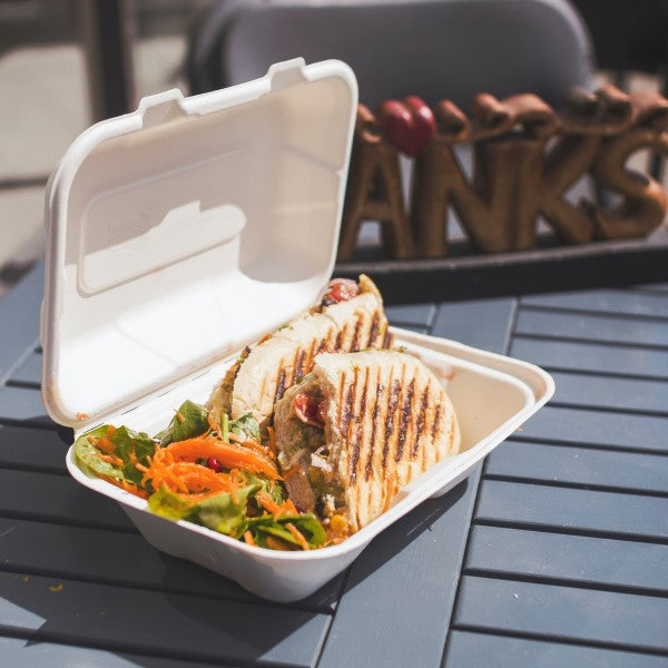 A takeout container with two grilled sandwiches and a fresh salad inside, placed on a blue table with a sign that says "Thanks" in the background.